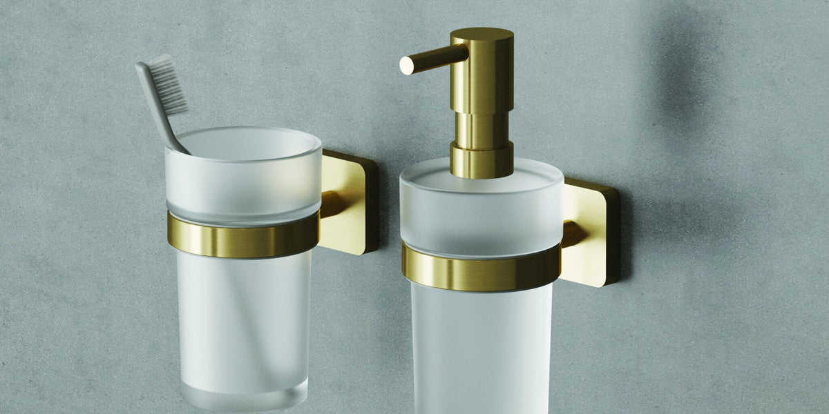 Wall-mounted soap dispenser in brushed gold beside a tumbler holder