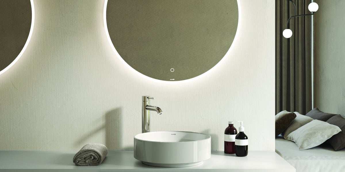 Round backlit mirrors over a vanity, with a bedroom beyond